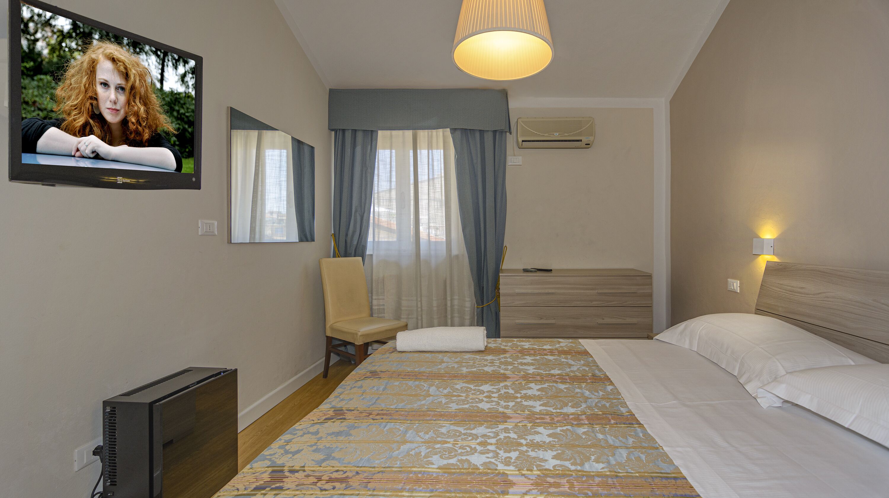 A hotel room with a bed, a chair, a TV, and a large window with curtains.