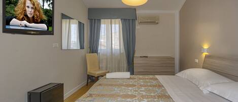Una habitación de hotel con cama, sillón, televisión y un ventanal con cortinas.