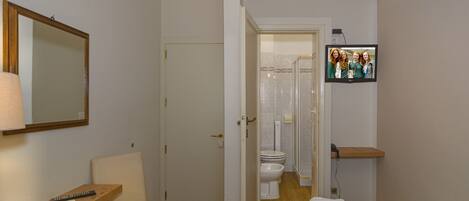 A hotel room with a wooden chair, a small table with a remote control, a bed with a patterned bedspread, a television mounted on the wall, and a door leading to a bathroom with a toilet and shower.