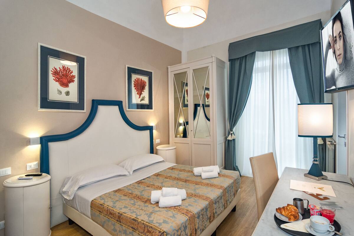 Ein Hotelzimmer mit Bett, Nachttisch, Lampe und einem Frühstückswagen mit Croissant, Saft und Kaffee.