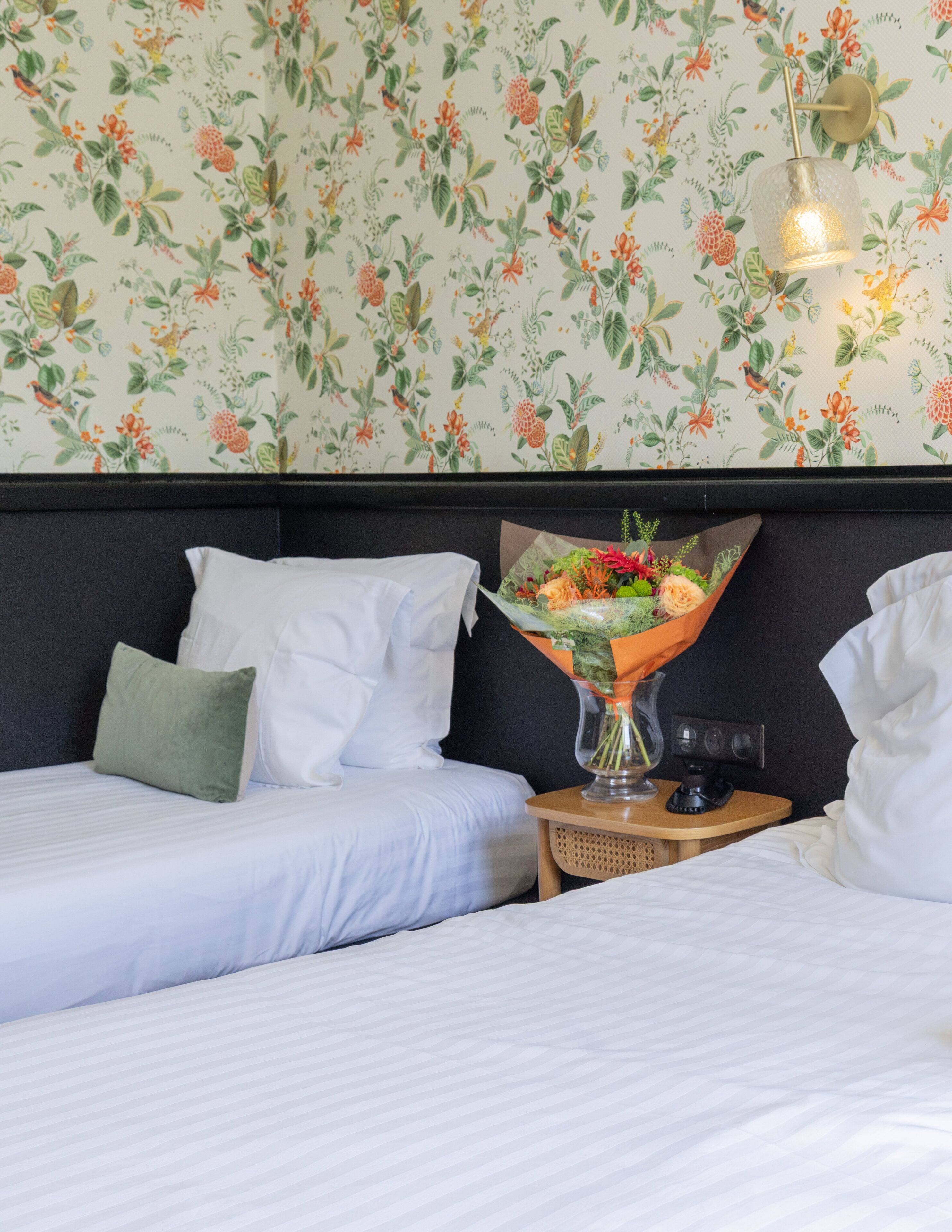 A hotel room with two beds, a nightstand, a lamp, and a floral wallpaper.