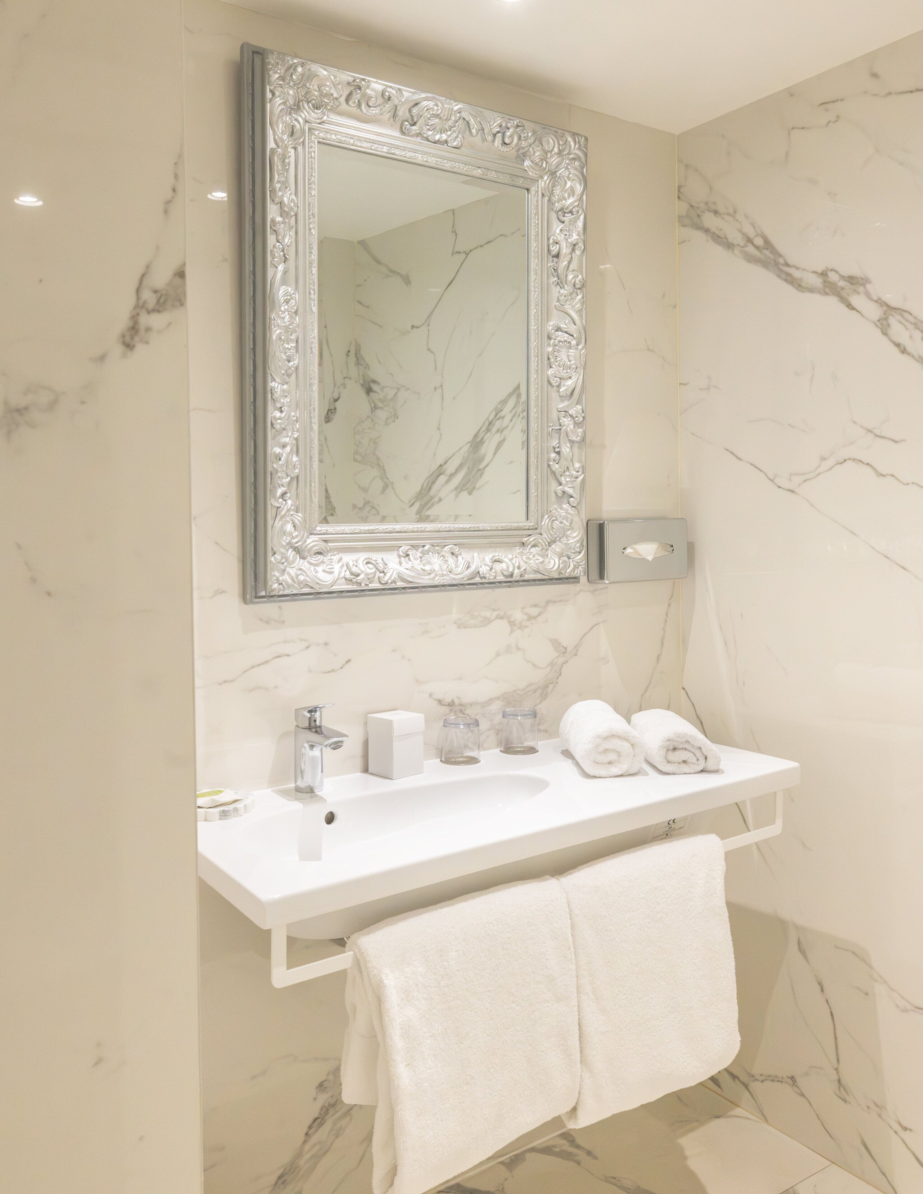 Une salle de bain avec un mur en marbre, un lavabo blanc, un miroir doté d’un cadre orné et deux serviettes posées sur une étagère.