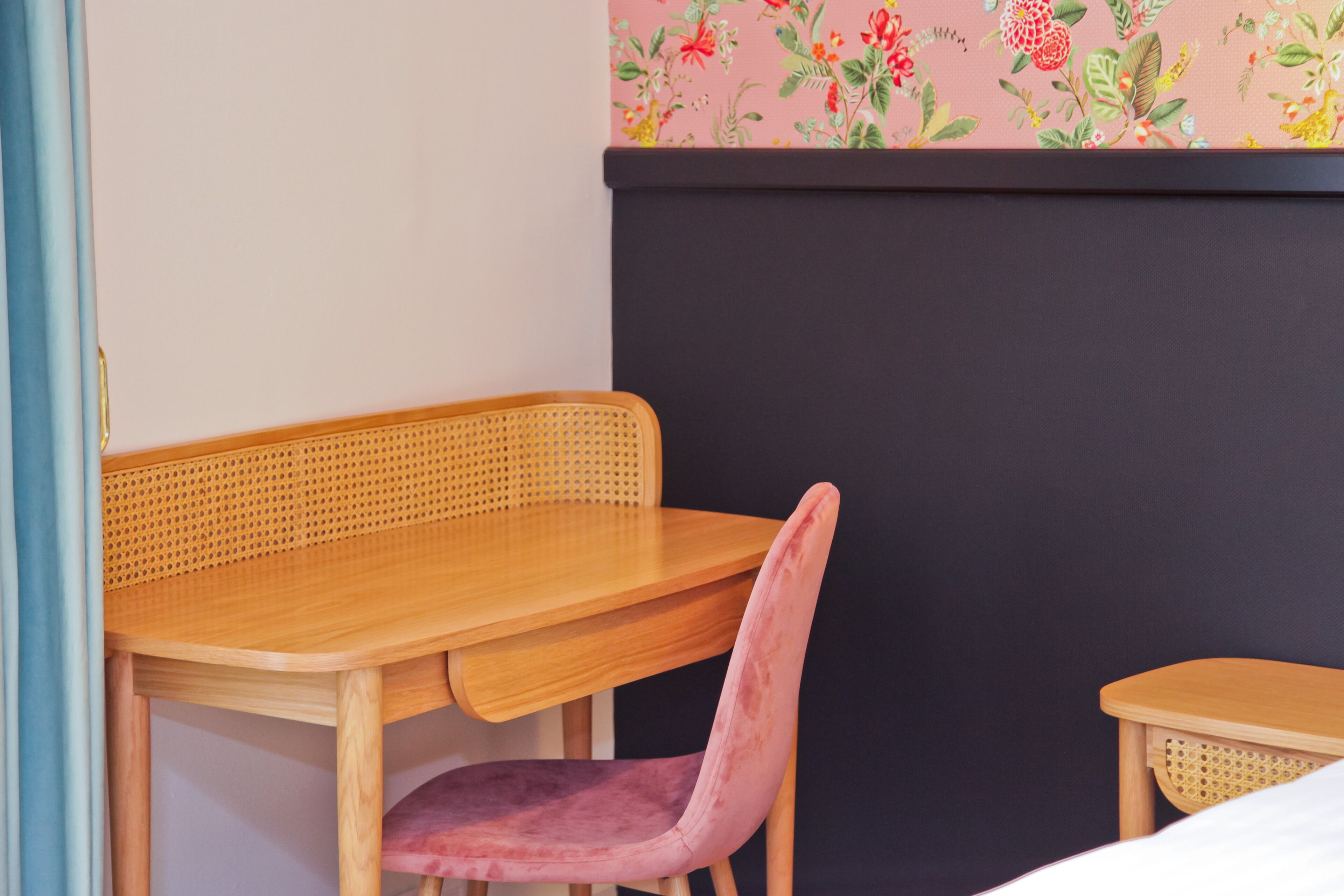 Een houten tafel met een geweven stoel, een klein bijzettafeltje en een donkere muur met bloemenbehang.