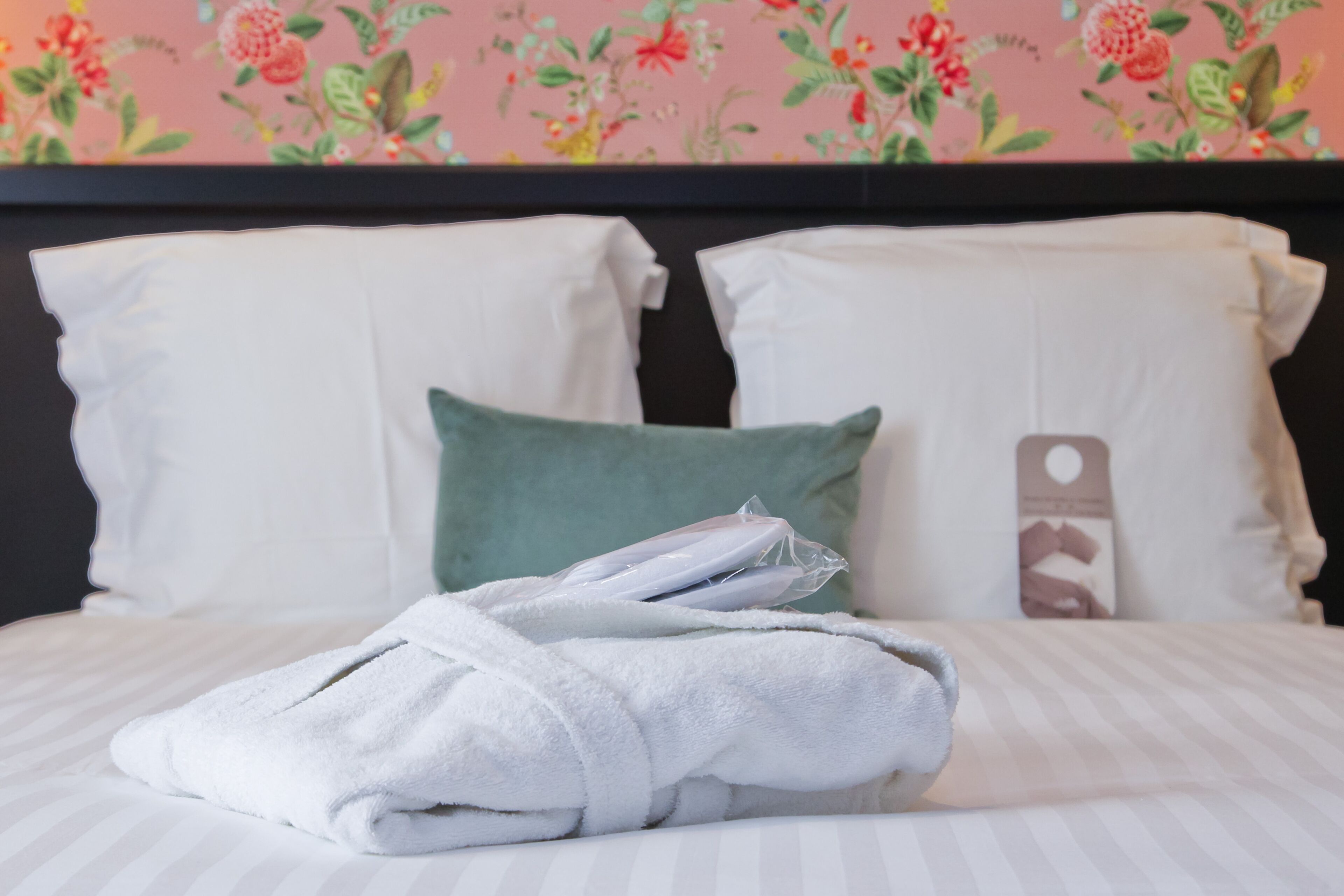 A neatly made bed with white and green pillows, a folded white towel, and a pillow with a phone case design.