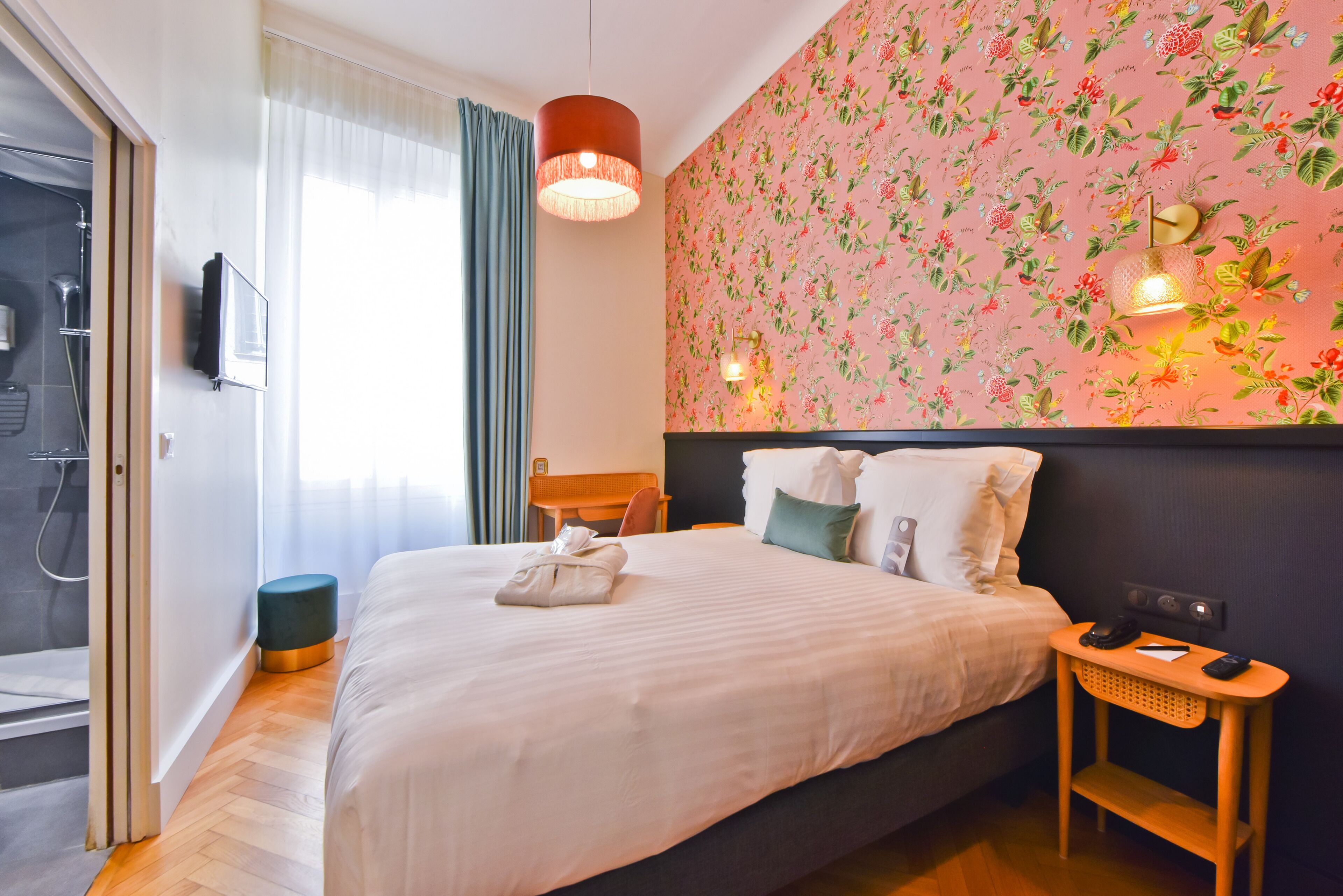 Habitación de hotel con cama, mesita de noche con teléfono, televisor montado en la pared, zona de ducha y papel tapiz con motivos florales.
