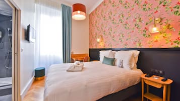 Habitación de hotel con cama, mesita de noche con teléfono, televisor montado en la pared, zona de ducha y papel tapiz con motivos florales.