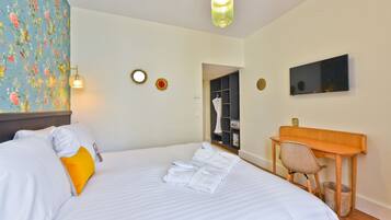 A neatly made bed with white and yellow pillows, a wooden desk with a chair, a mirror, and a television on the wall.