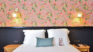 Una habitación de hotel con papel tapiz floral, una cama con almohadas blancas y turquesa, dos mesitas de noche con lámparas, y un mueble auxiliar con un teléfono y un control remoto.