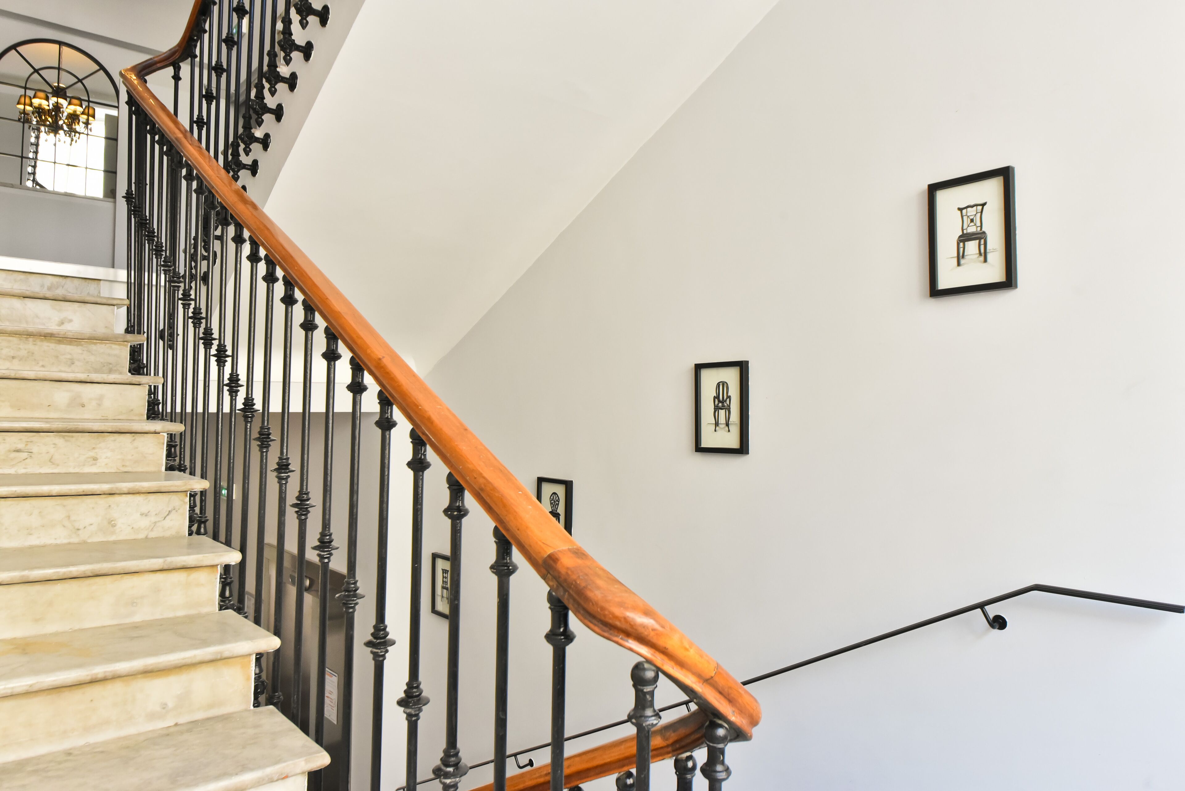 Eine Treppe mit hölzernem Geländer und schwarzen Metallspindeln, die zu einer Zwischenebene mit gerahmten Kunstwerken an der Wand führt.