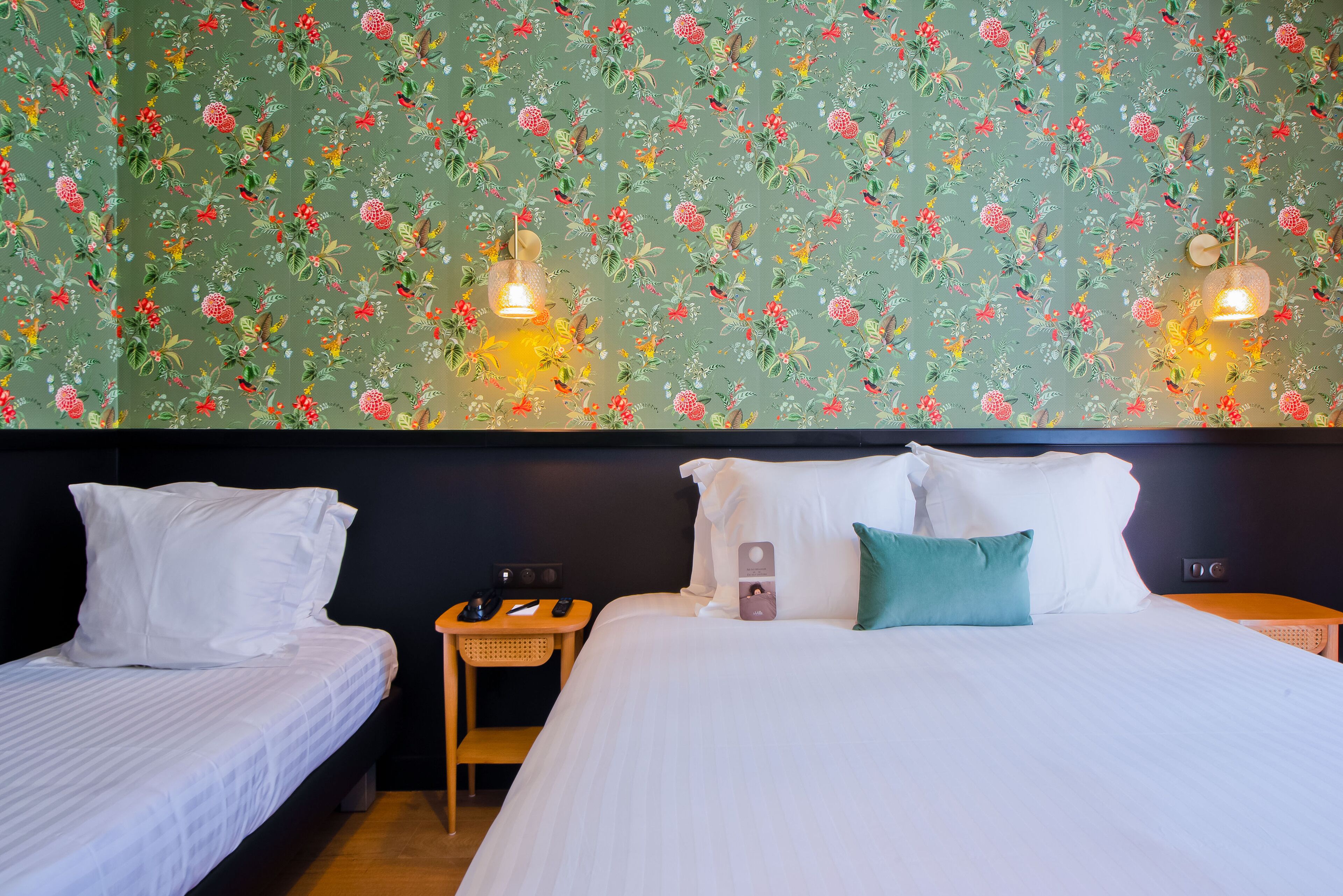 Habitación de hotel con dos camas, una mesita de noche de madera, un papel tapiz floral y dos lámparas de pared.