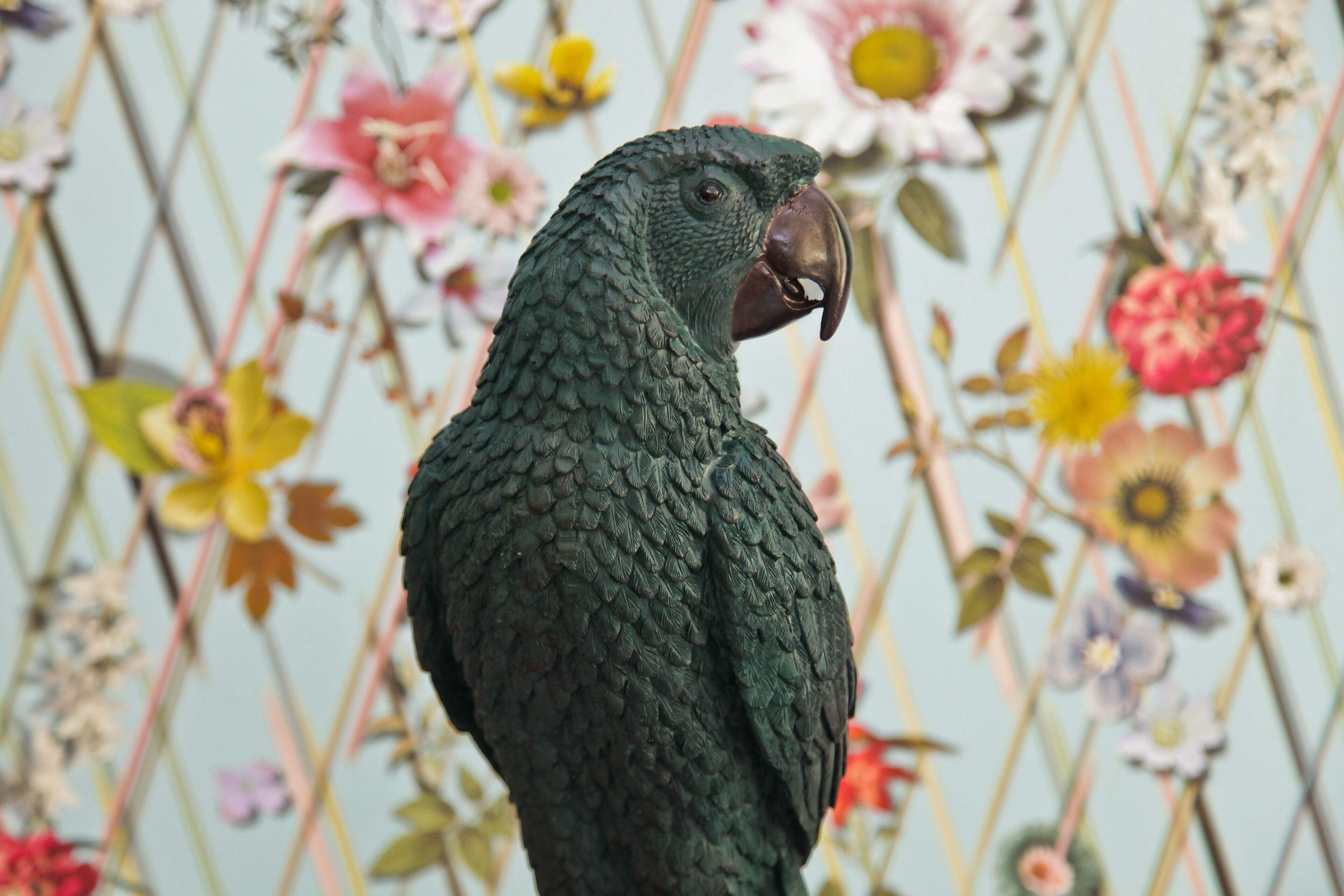 A parrot with a dark green plumage and a brown beak, standing against a backdrop of various flowers.