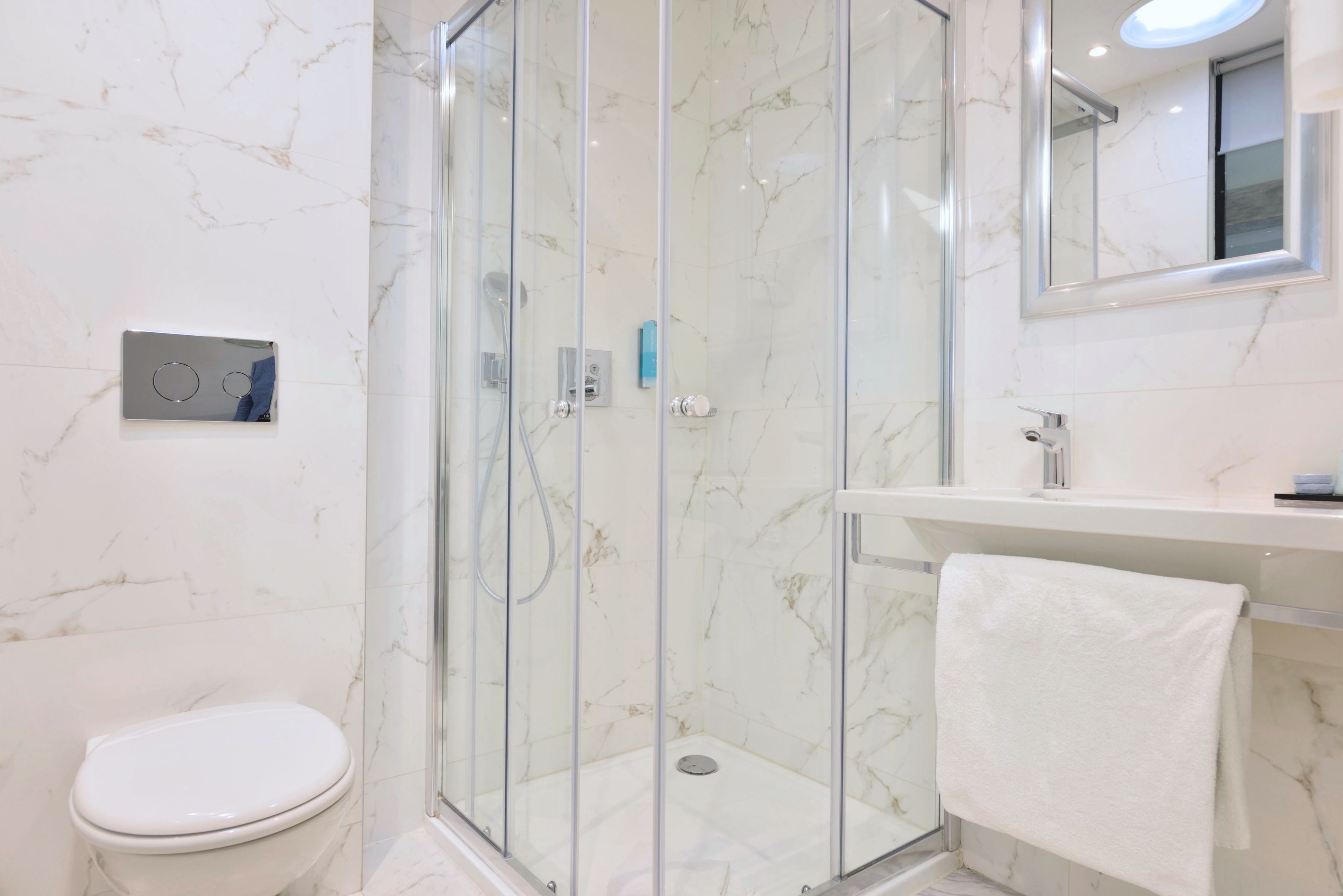 A modern bathroom with a glass-enclosed shower, a white toilet, a marble-tiled wall, and a sink with a towel hanging on the side.