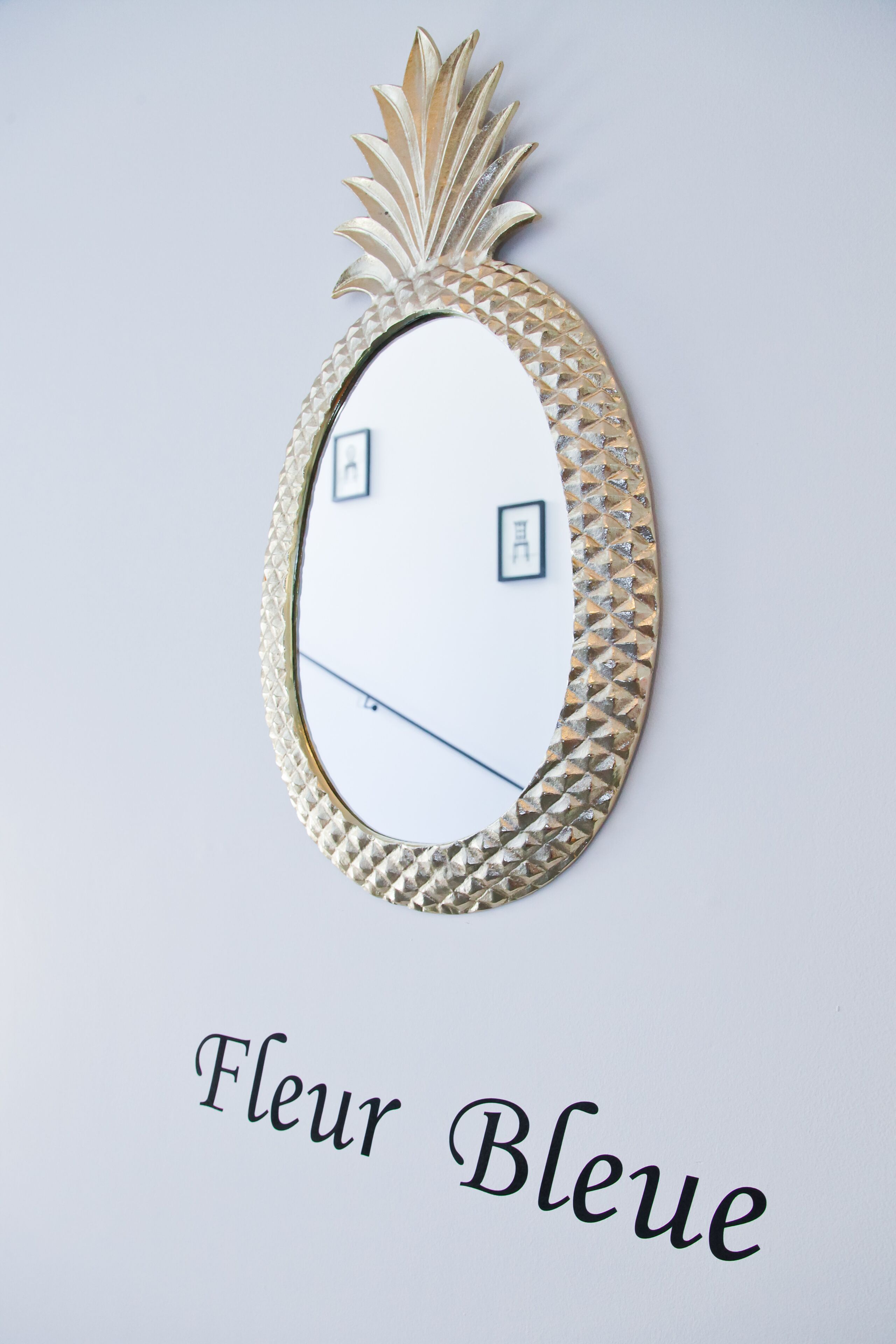 Un espejo fijado a la pared con forma de piña en la parte superior y el texto "Fleur Bleue" debajo.