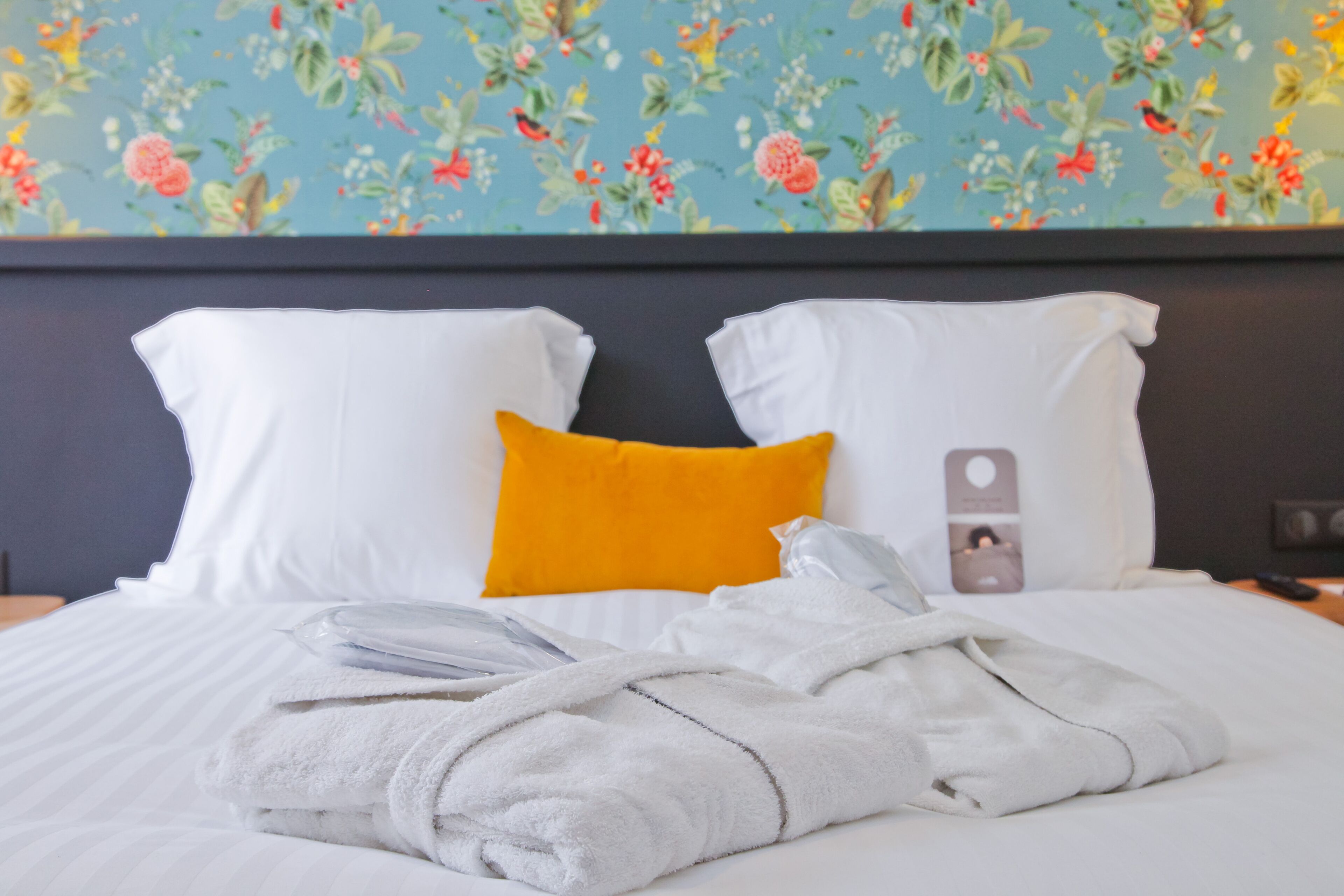 A neatly made bed with white and yellow pillows, a folded white towel, and a white robe on a bed with a floral wallpaper background.