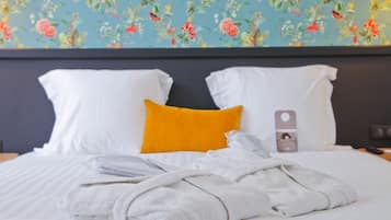 A neatly made bed with white and yellow pillows, a folded white towel, and a white robe on a bed with a floral wallpaper background.
