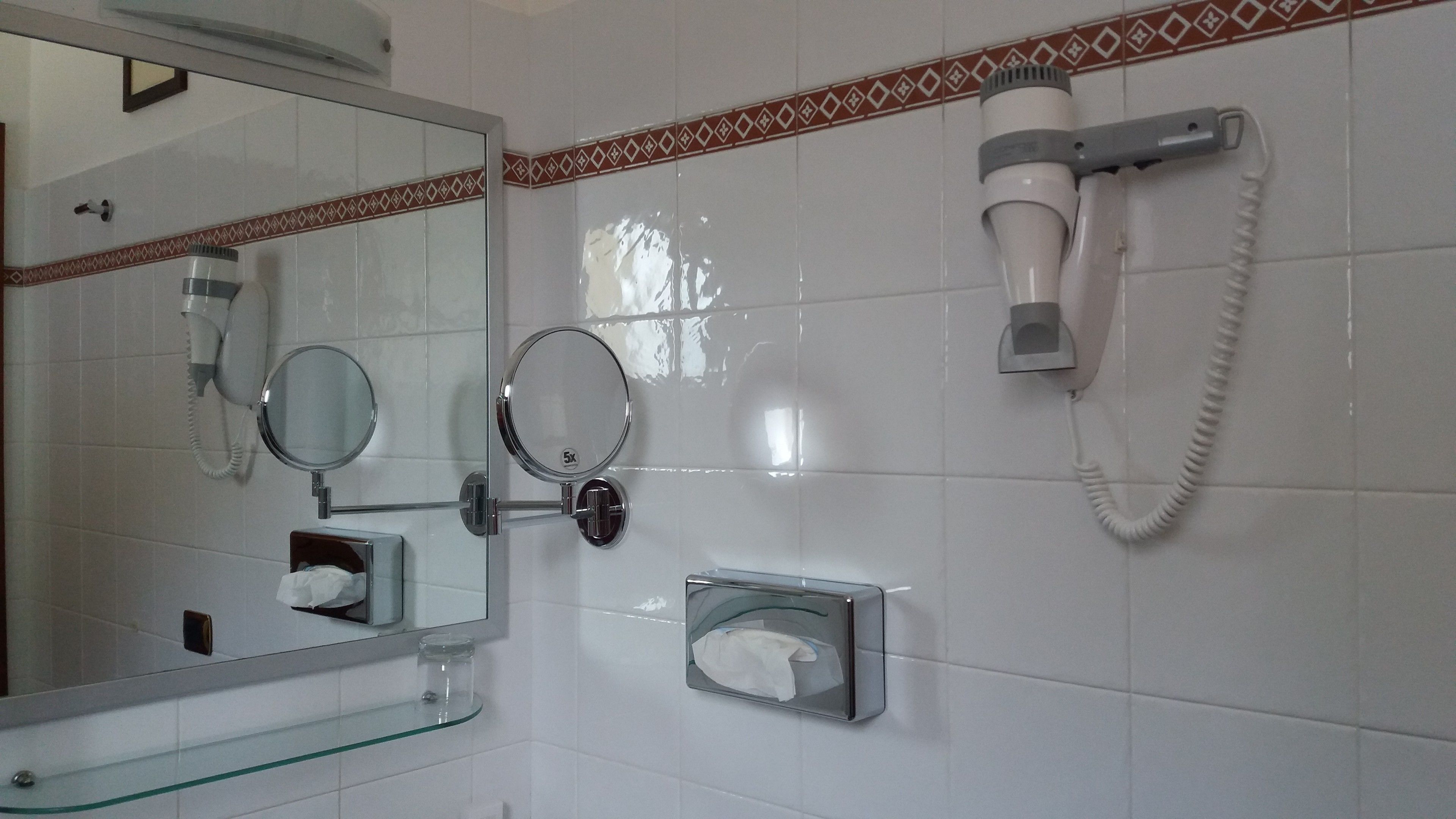 Un baño con espejo, dos secadores de pelo, dispensador de pañuelos de papel y repisa de vidrio.