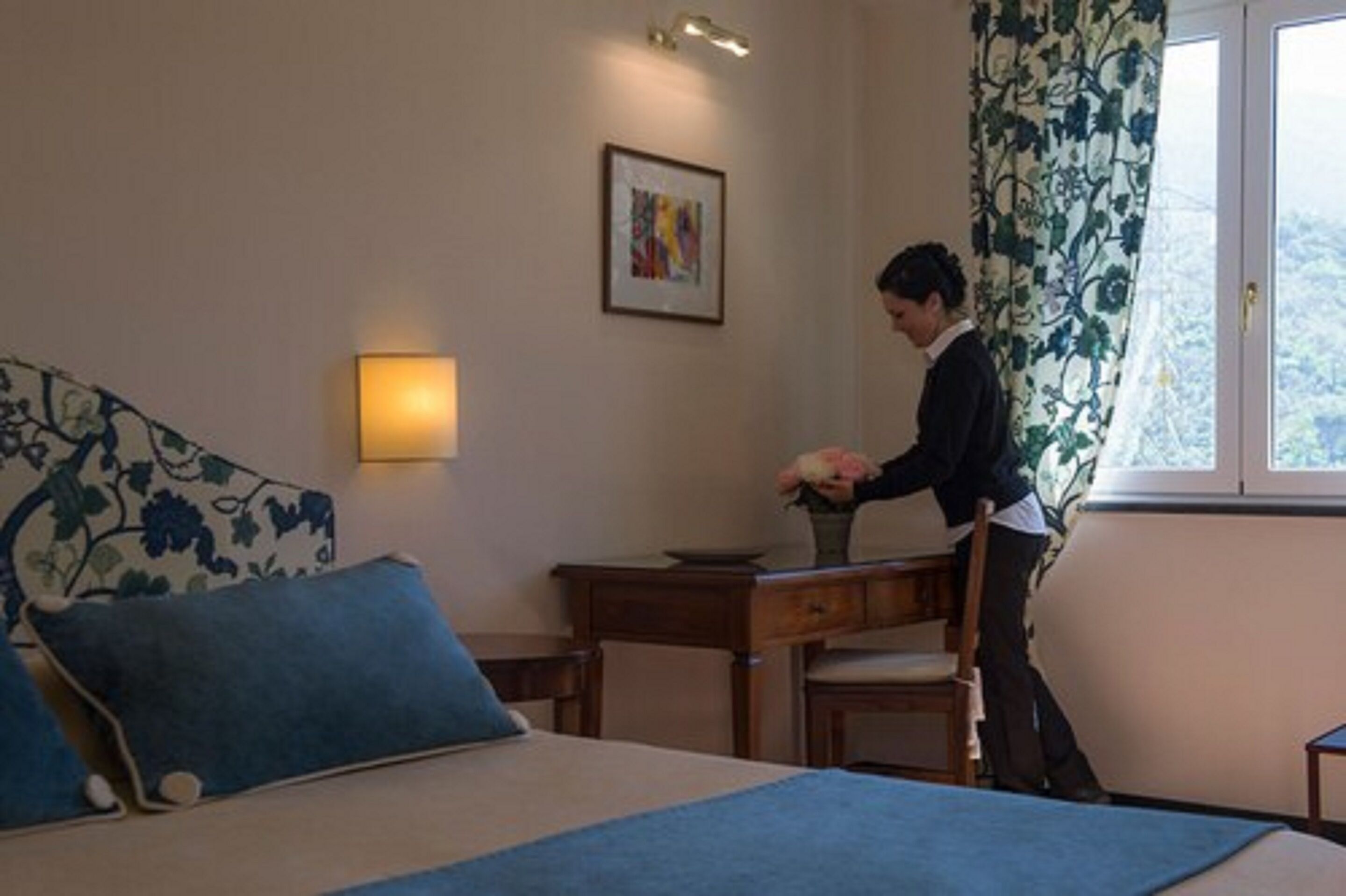 Une chambre d’hôtel avec un lit, un bureau, une chaise, un vase de fleurs, un tableau encadré au mur et une fenêtre avec des rideaux.