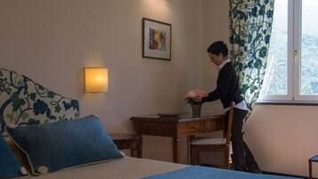 Une chambre d’hôtel avec un lit, un bureau, une chaise, un vase de fleurs, un tableau encadré au mur et une fenêtre avec des rideaux.
