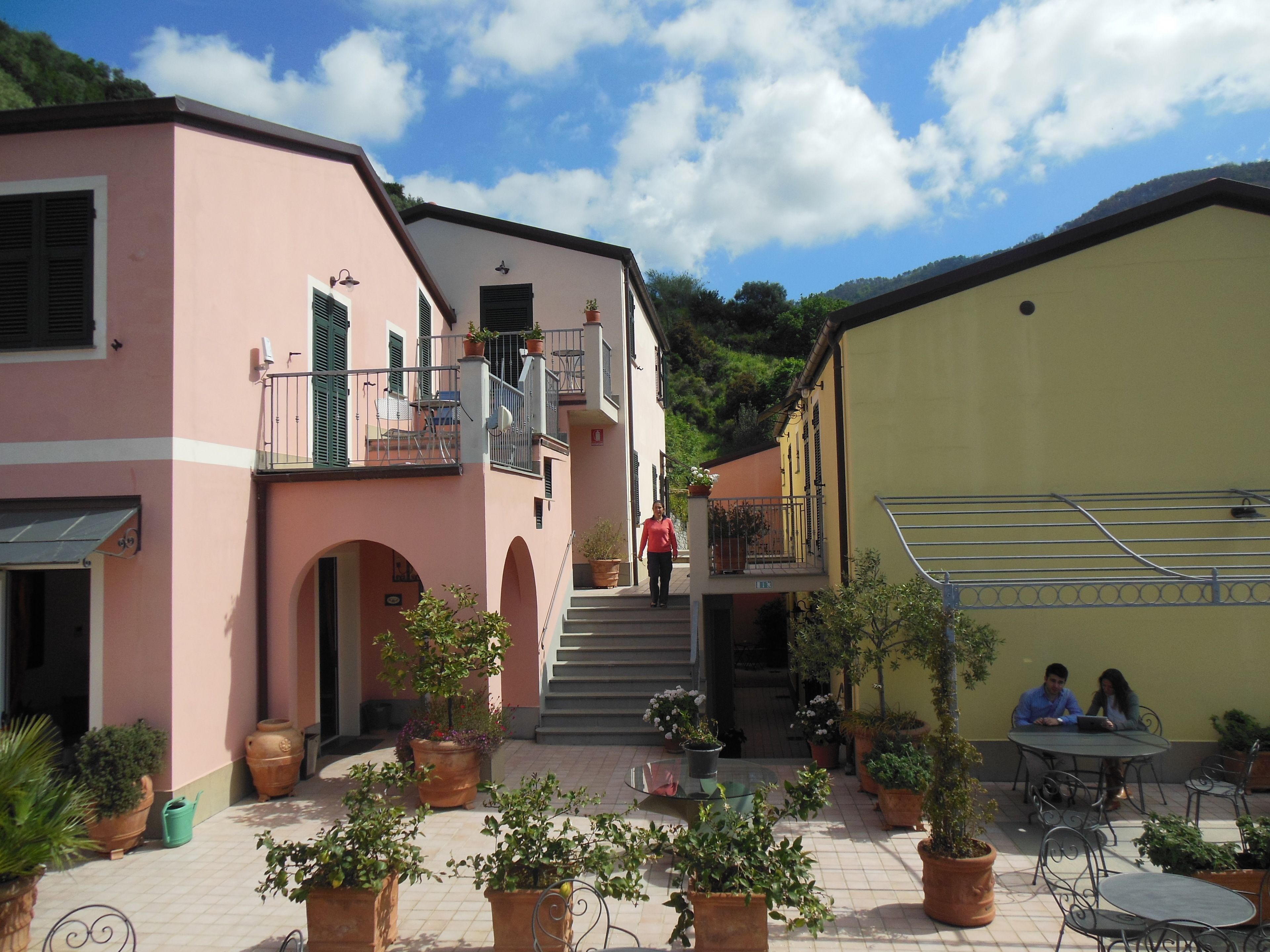 A courtyard with potted plants, a staircase leading to a balcony, and a table with two people sitting.