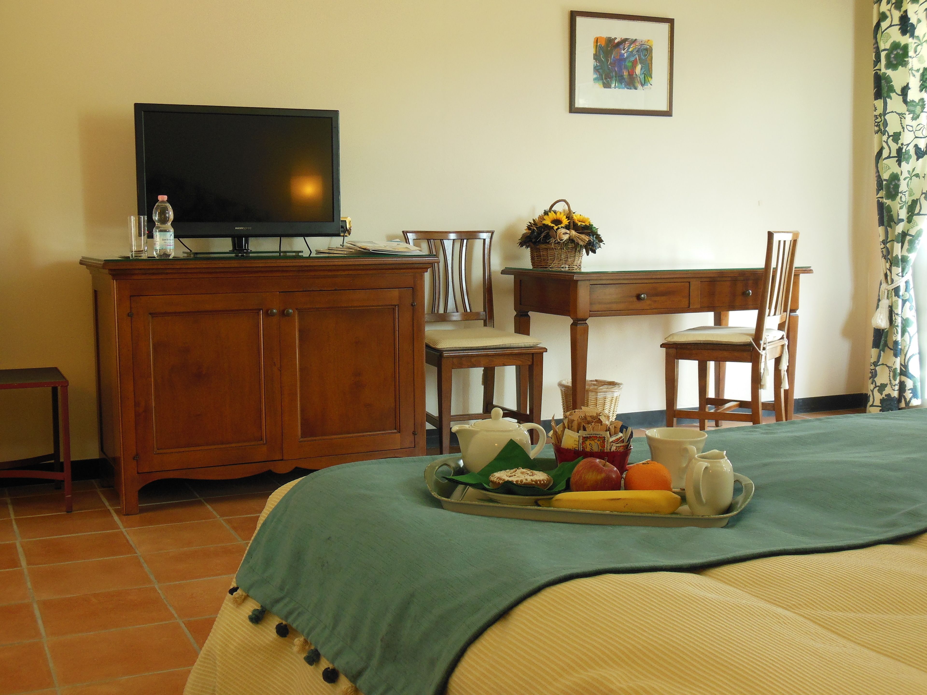 A hotel room with a wooden desk, a television, a dining table with a fruit tray, and a bed with a green bedspread.