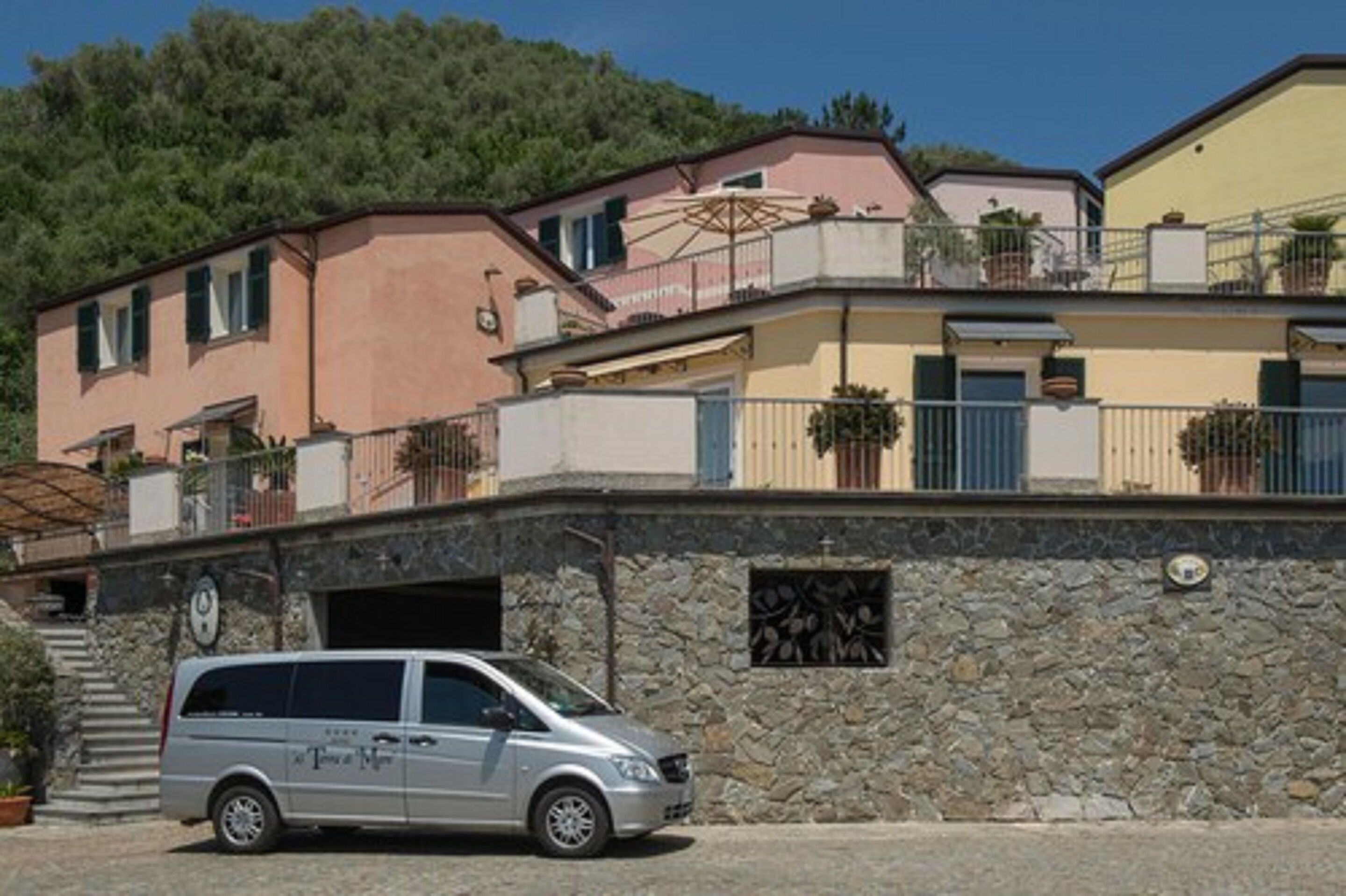 Una hilera de edificios color terracota con balcones y plantas en macetas, un muro de piedra con una reja de metal, y una furgoneta plateada estacionada.