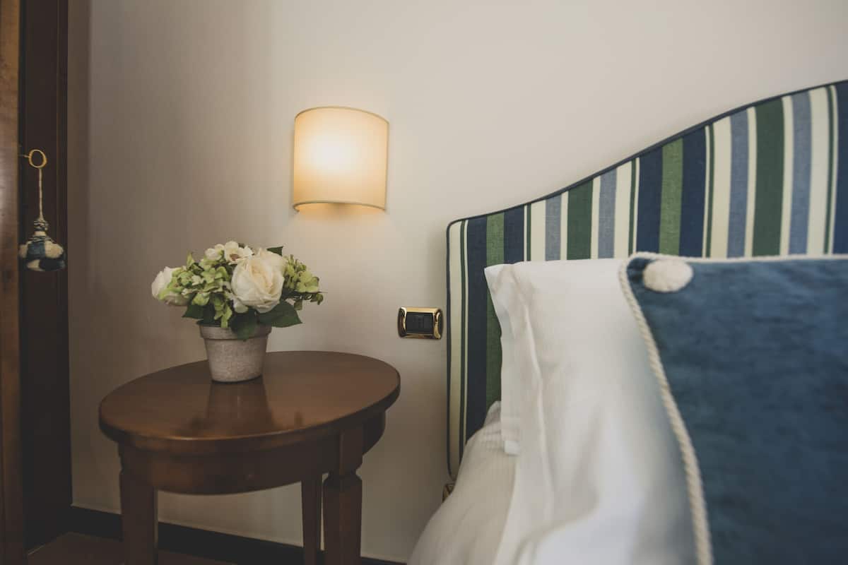 A hotel room with a bed, a bedside table with a flower vase, a wall-mounted light, and a door with a keyhole.