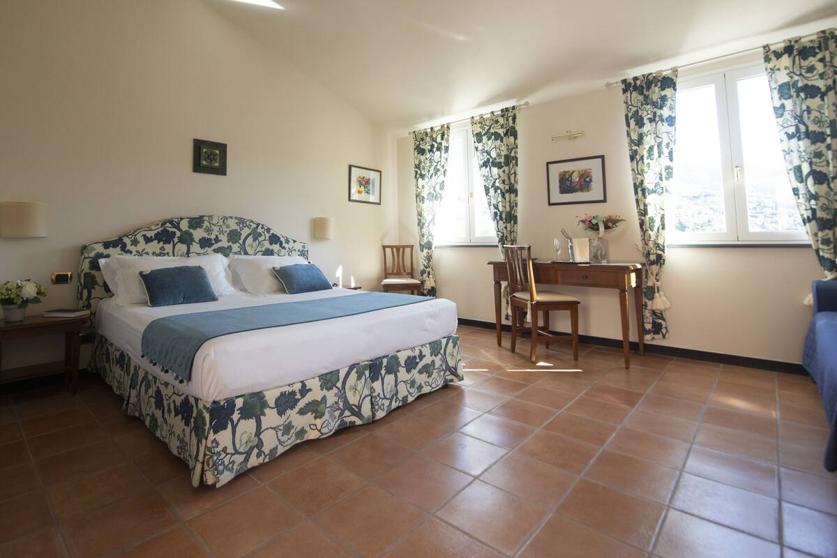 A bedroom with a bed, a desk, and a chair. There are two windows with curtains.