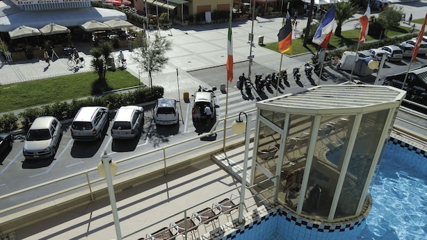 A swimming pool with a person swimming, surrounded by a parking area with cars, a building with a sign, and a view of the sea. - Hotel Residence Villa Jolanda (Camaiore)