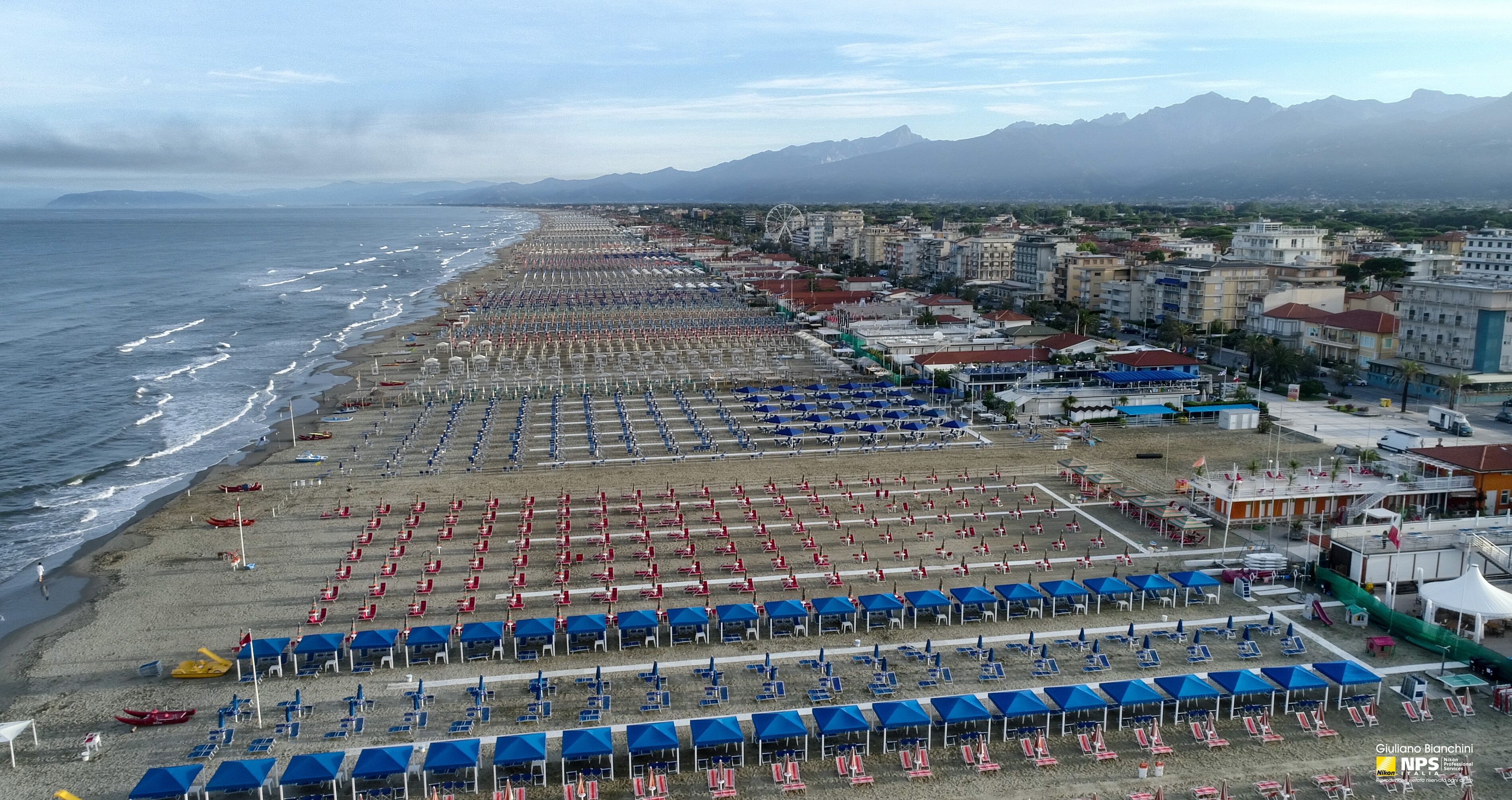 A beach with rows of blue and red umbrellas and chairs, a Ferris wheel, and buildings in the background.