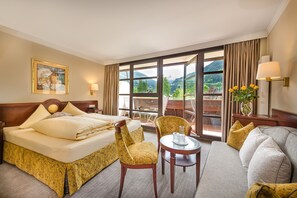 Habitación de hotel con una cama grande, dos sillas, una mesita y vistas a las montañas a través de amplios ventanales.