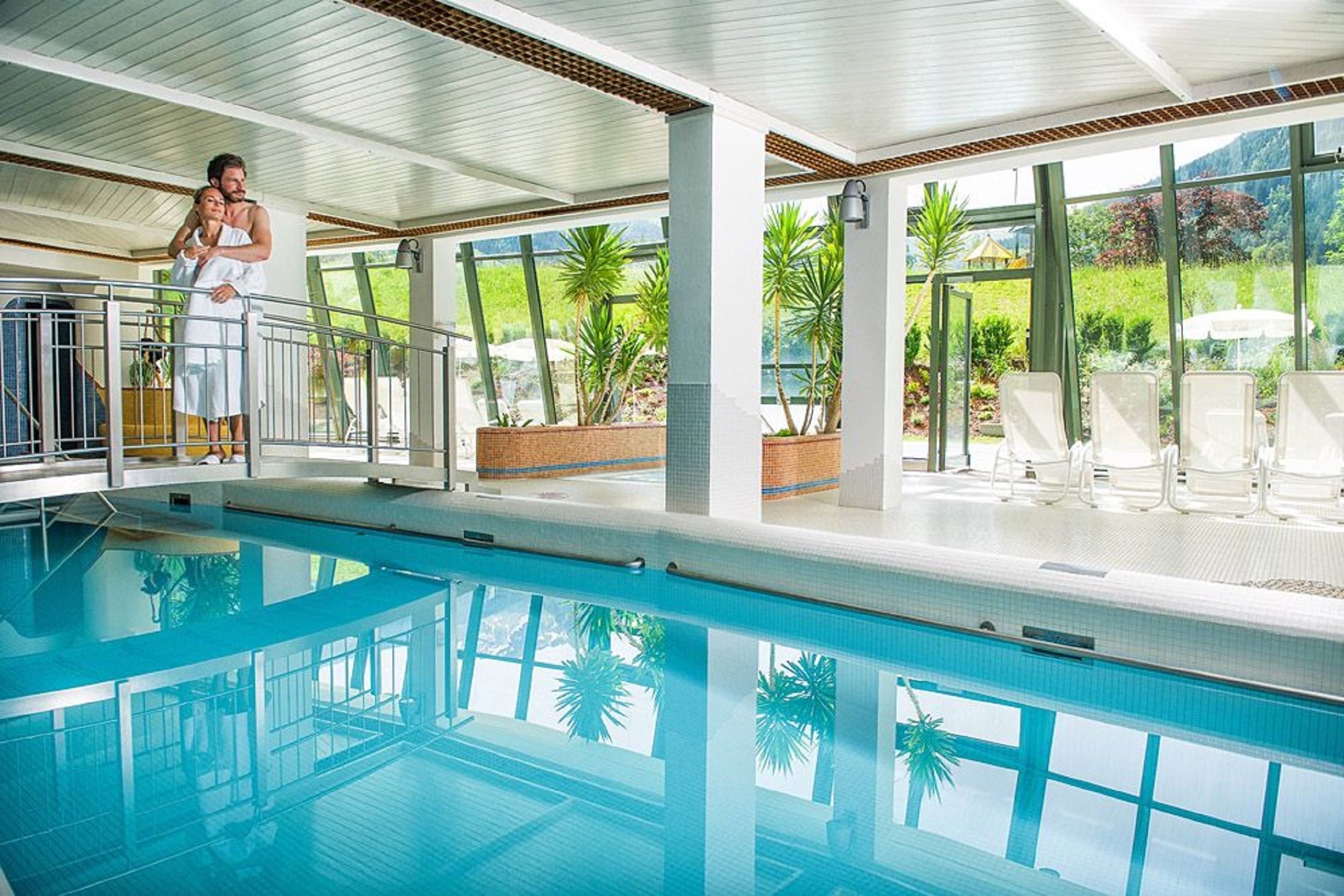 Una piscina interior con paredes de vidrio transparente, suelo de baldosas y vistas a la vegetación exterior.