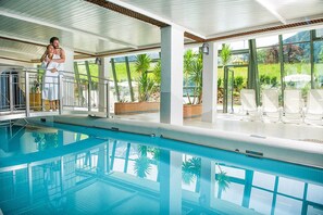 Una piscina interior con paredes de vidrio transparente, suelo de baldosas y vistas a la vegetación exterior.