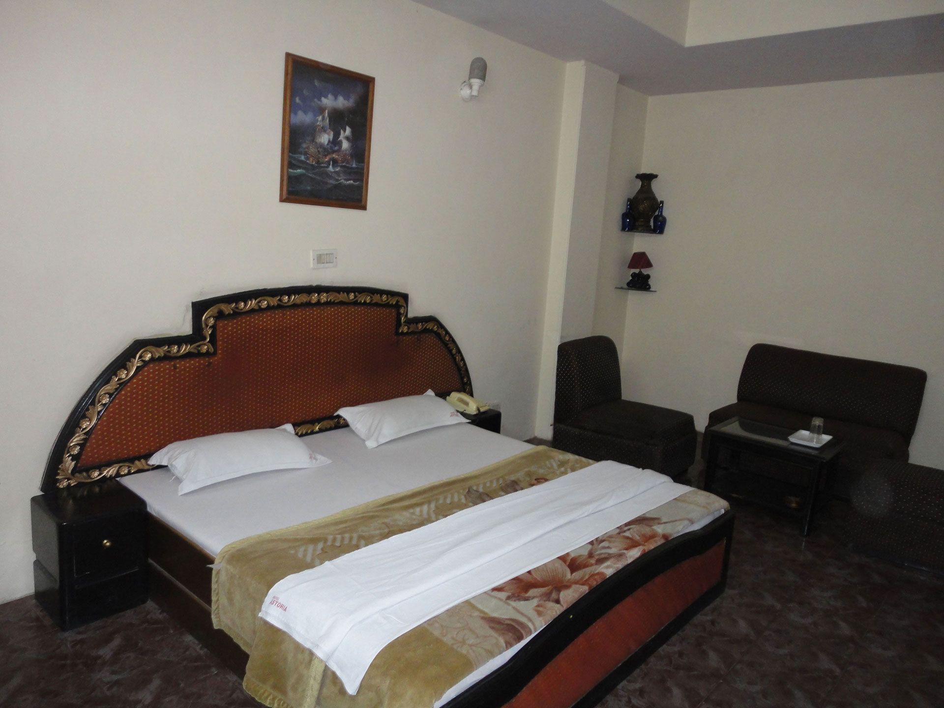 A hotel room with a large bed, a chair, a small table, and a wall-mounted lamp.