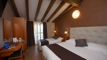 A hotel room with two beds, a wooden desk with a chair, a small table, and a window with curtains.