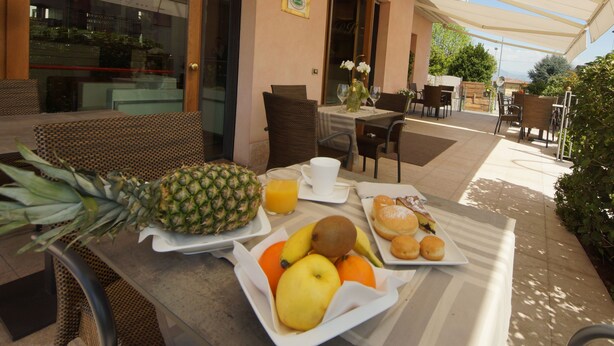 A breakfast setup with a pineapple, oranges, kiwi, and a plate of pastries on a table outside a building with wicker furniture and a covered patio.