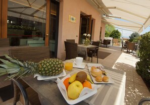 A breakfast setup with a pineapple, oranges, kiwi, and a plate of pastries on a table outside a building with wicker furniture and a covered patio. - Hotel Clarin 14 by Dori (Peschiera del Garda)