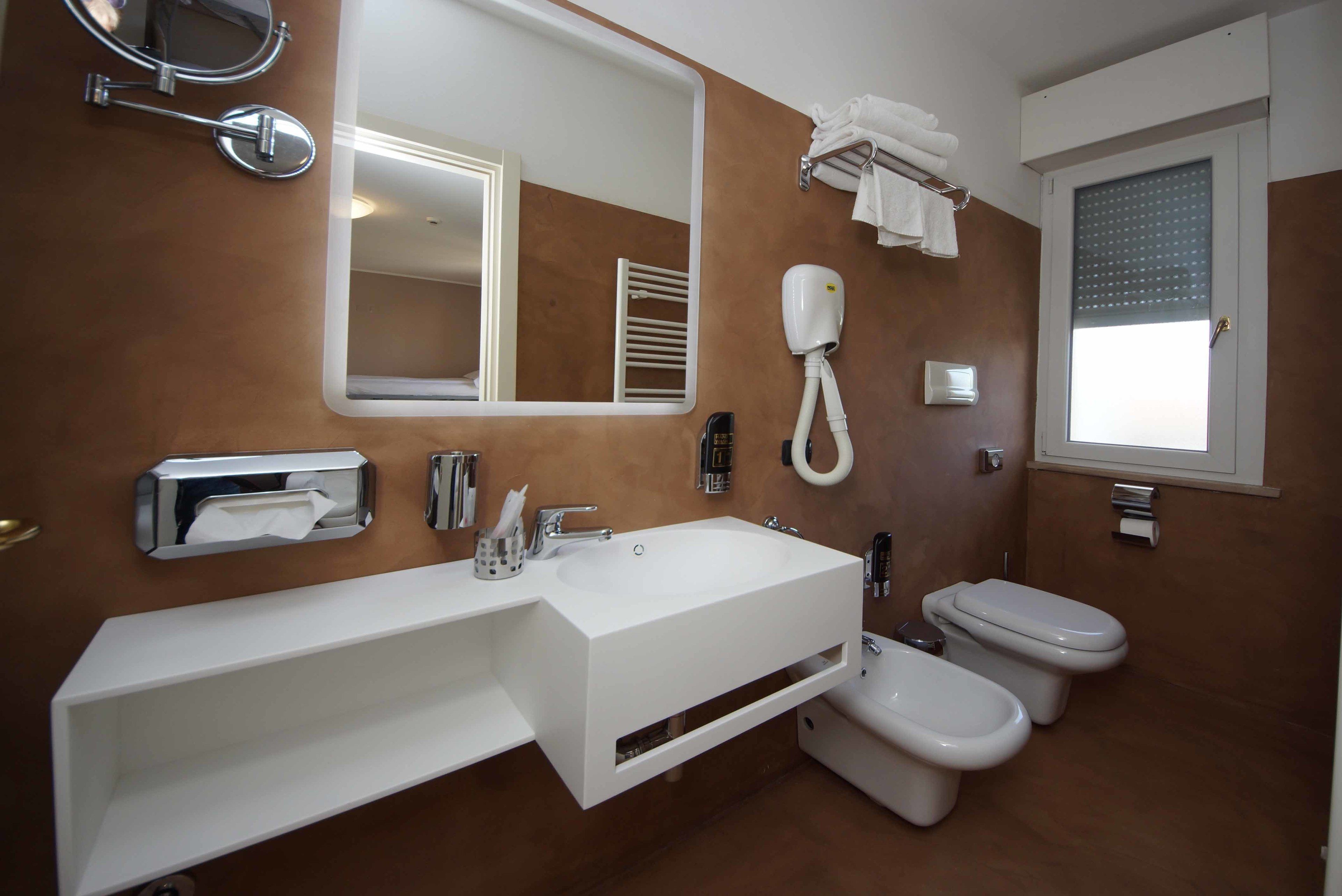 Un bagno moderno con un lavandino bianco, uno specchio grande, un portasciuga, un water e una finestra con persiane.
