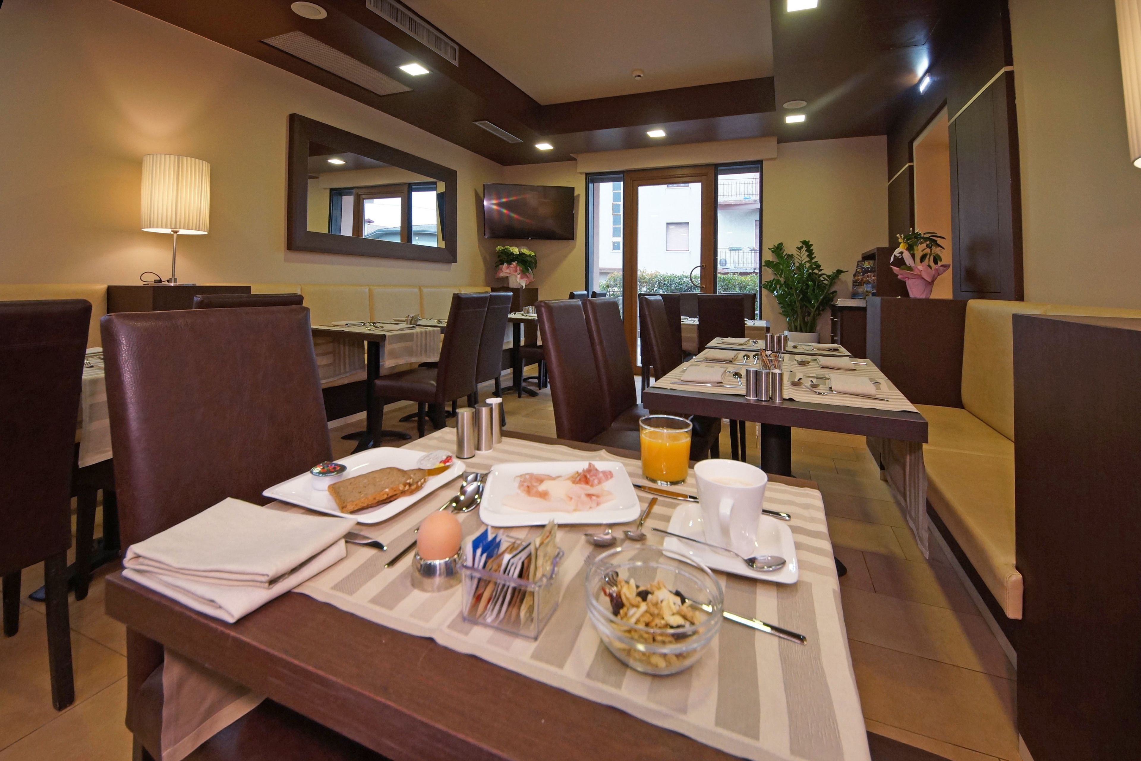 Uma sala de jantar de hotel com mesas preparadas para o pequeno-almoço, incluindo pratos com comida, chávenas e uma taça de cereais.
