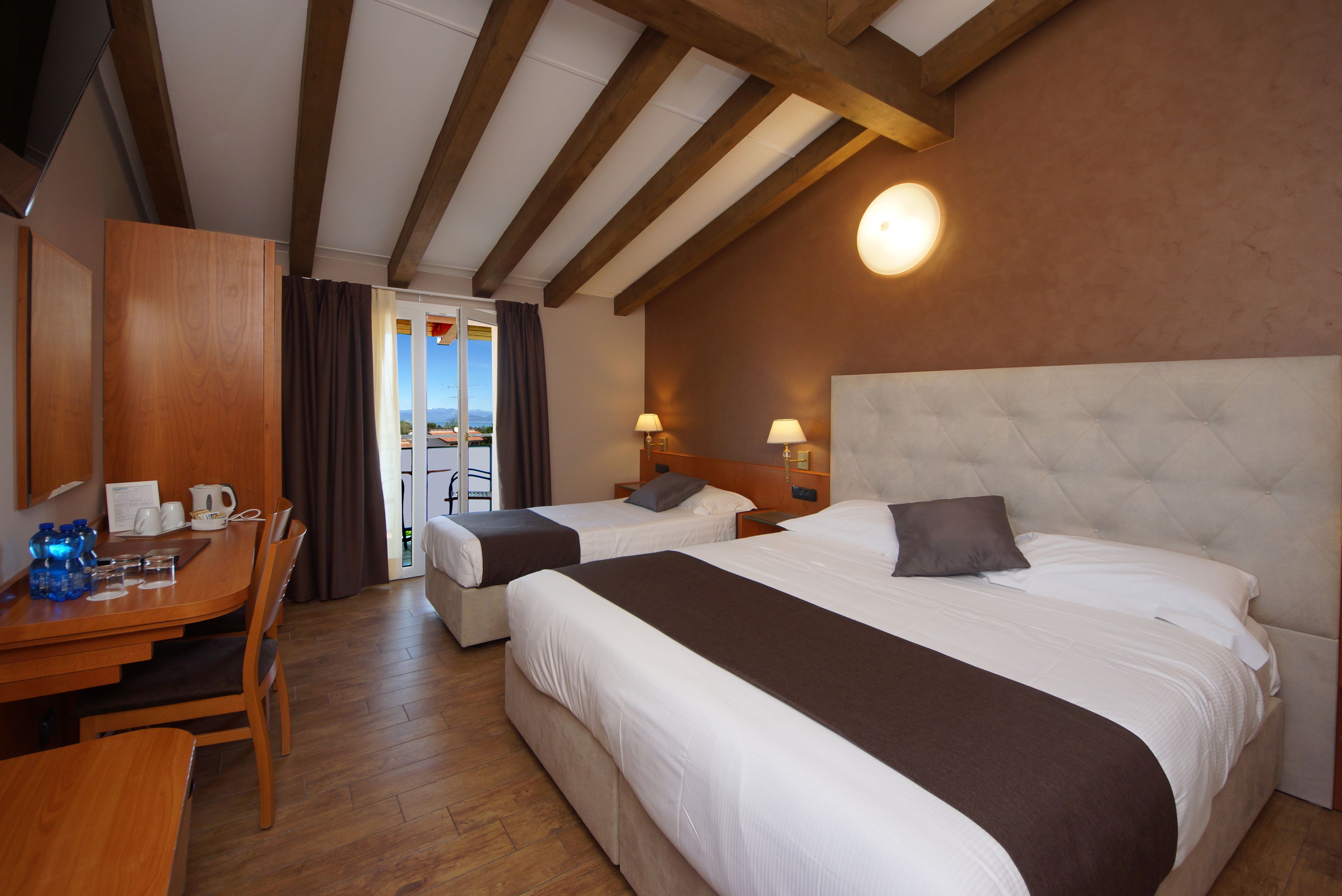 A hotel room with two beds, a wooden desk with a chair, a table with a kettle and cups, and a view of the outdoors.