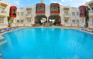 A large swimming pool with a clear blue water surface, surrounded by lounge chairs and a building with balconies.