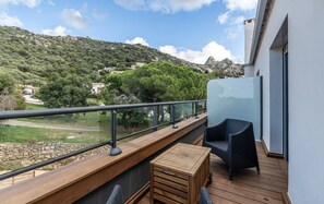 A balcony with a wooden table and chair, a glass railing, and a view of a green landscape with trees and a mountain in the background. - A Casa Di Mà (Lumio)