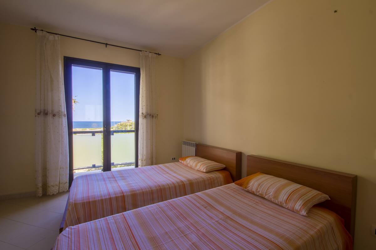 A room with two beds, a view of the sea through a large window, and a balcony.