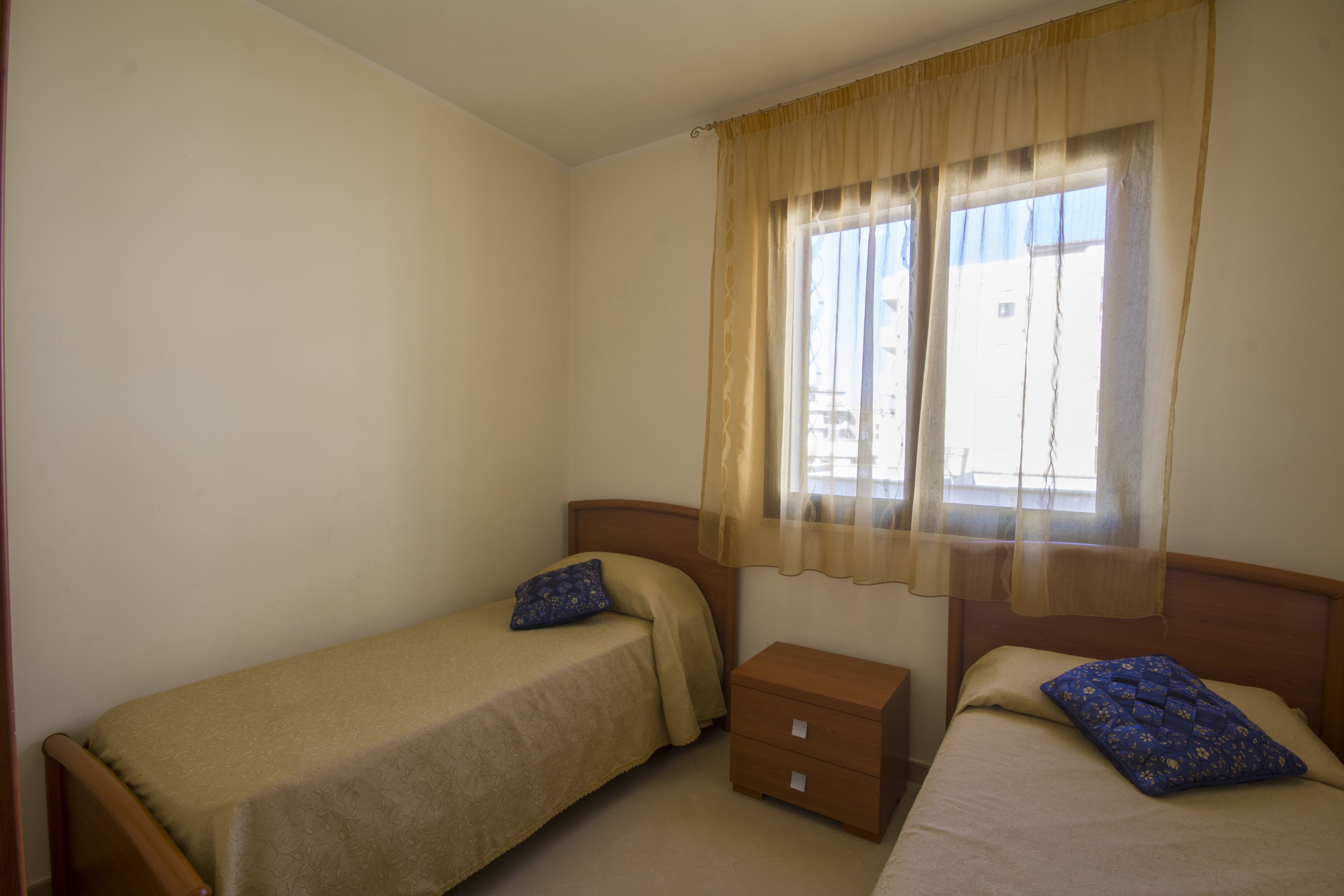 Una habitación pequeña y sencilla con dos camas individuales, una mesita de noche de madera y una ventana con cortina ligera.