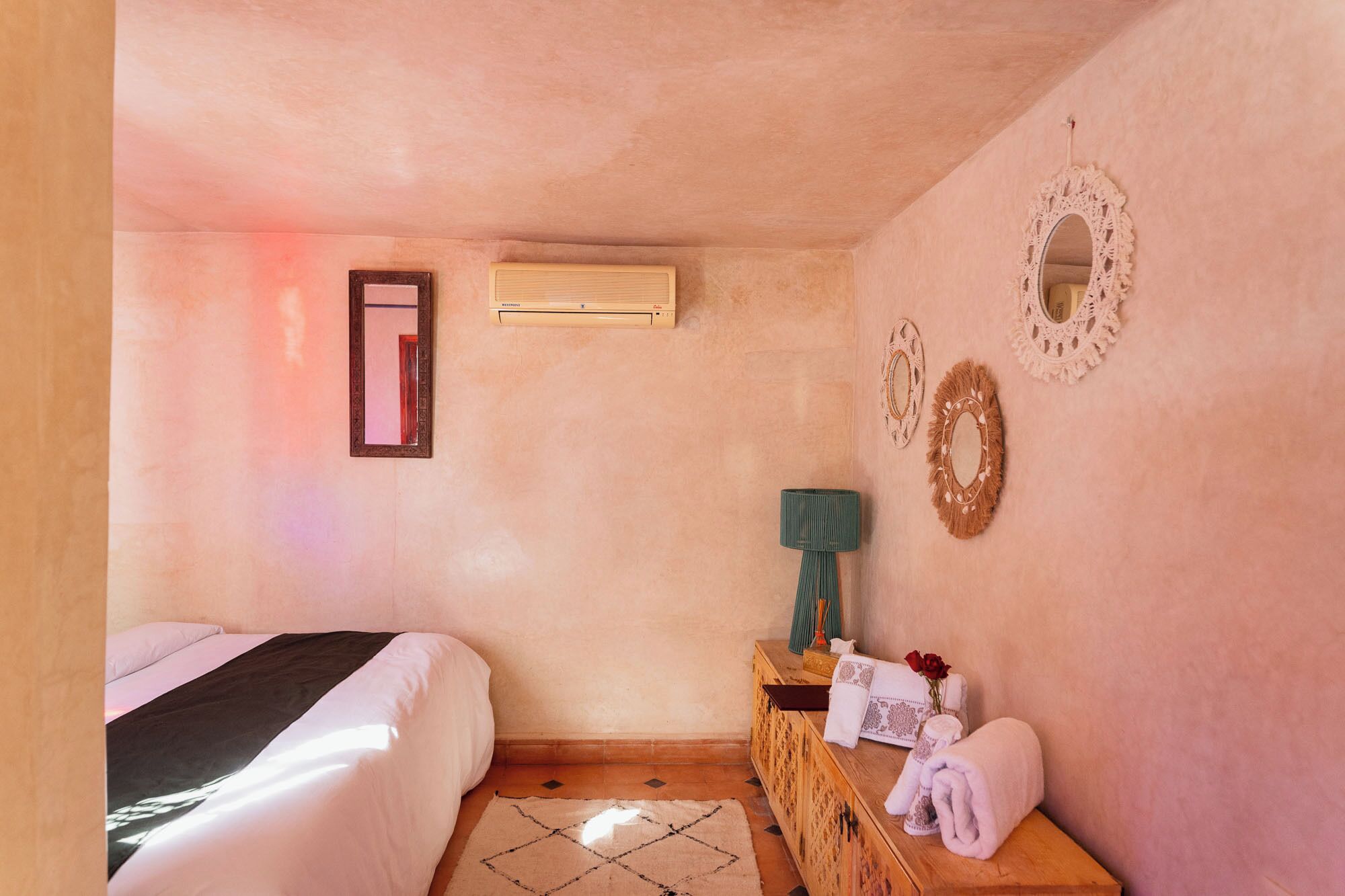 A bedroom with a bed, an air conditioner, a mirror, a lamp, and a wooden side table with towels and a vase of flowers.