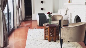 A cozy living room with a white brick fireplace, wooden beams on the ceiling, a large window with curtains, a sofa, a coffee table, and a rug. - Riad Twilight (Marrakech)