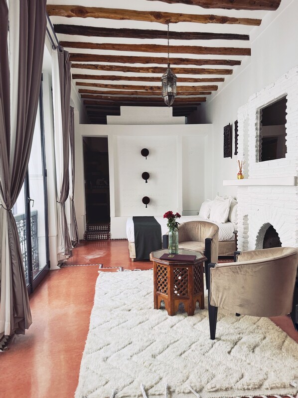 A cozy living room with a white brick fireplace, wooden beams on the ceiling, a large window with curtains, a sofa, a coffee table, and a rug.