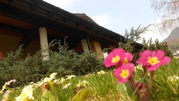 A building with a sloped roof and wooden beams, surrounded by lush greenery and flowers.