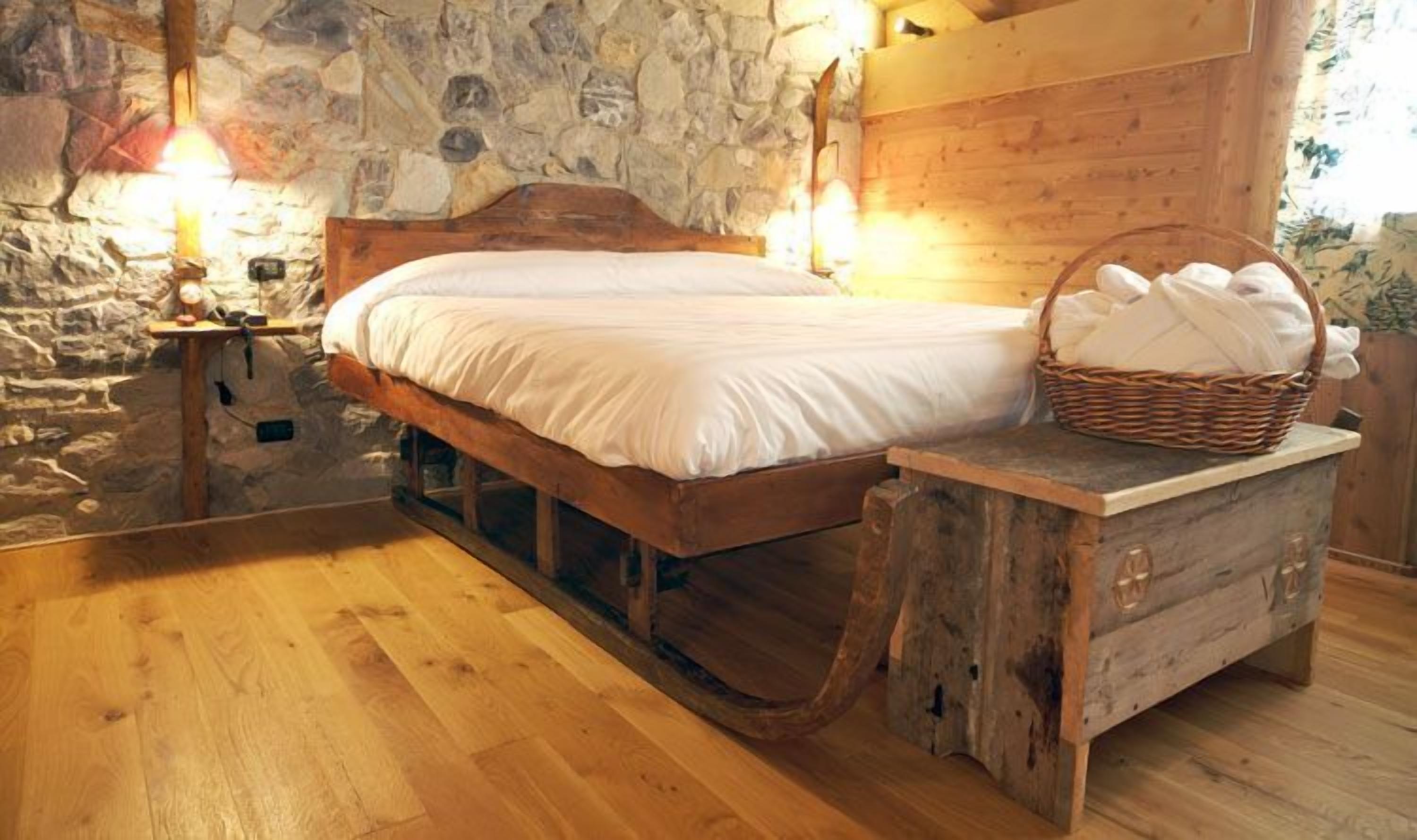 A rustic wooden bed with a stone wall backdrop, a nightstand with a lamp, and a basket of towels.