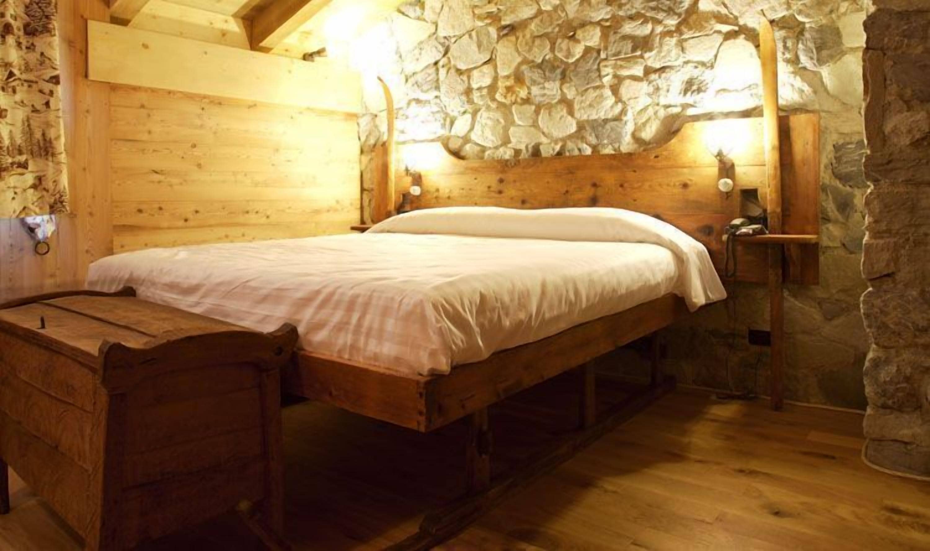 Una habitación rústica de madera con pared de piedra, una cama de madera, un banco y una mesita de noche con lámpara.