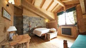 Un dormitorio de cabaña de madera con pared de piedra, cama, una mesita, lámpara y ventana con cortinas.